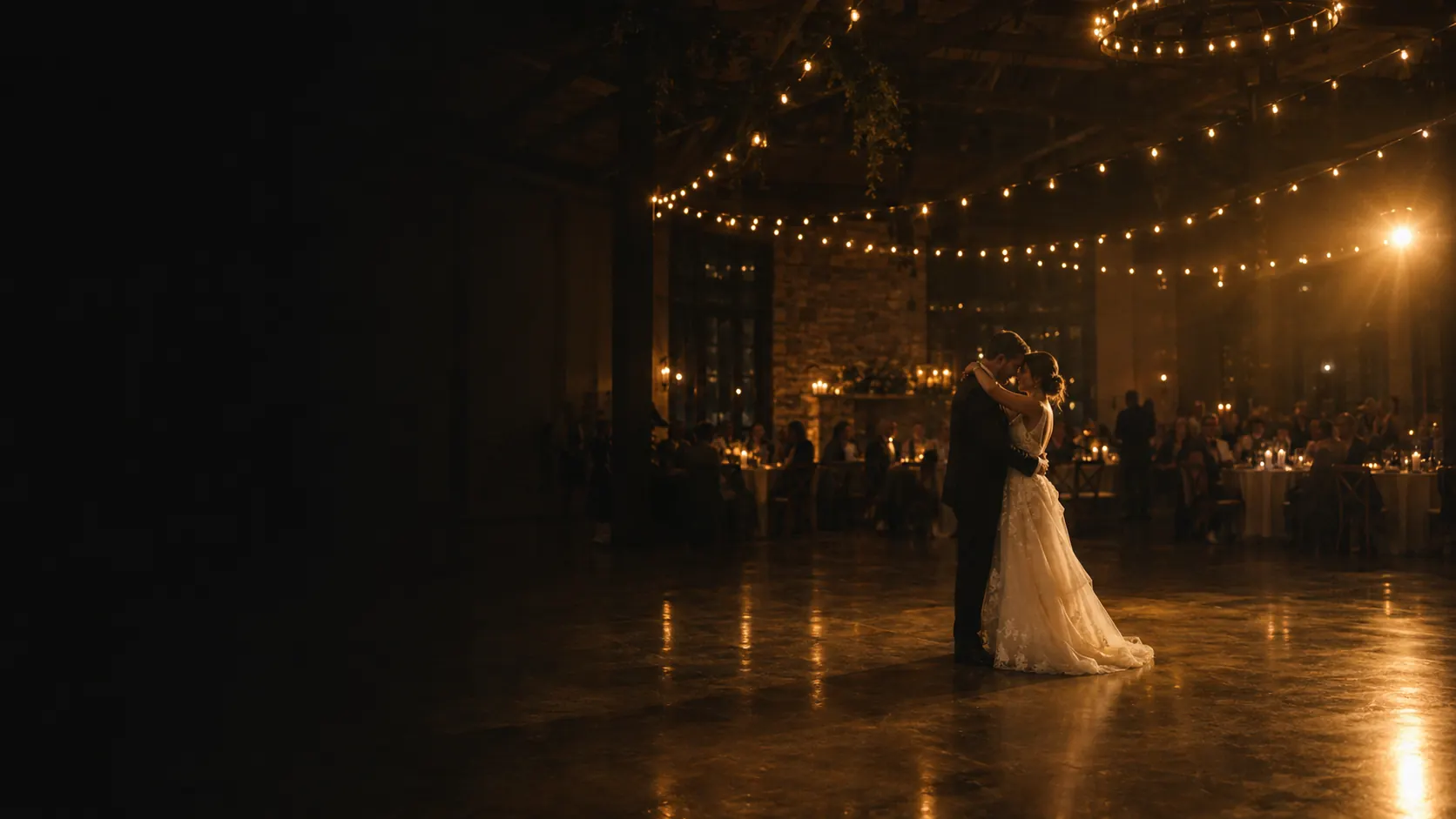 A couple sharing their first dance under warm string lights in a dark reception hall